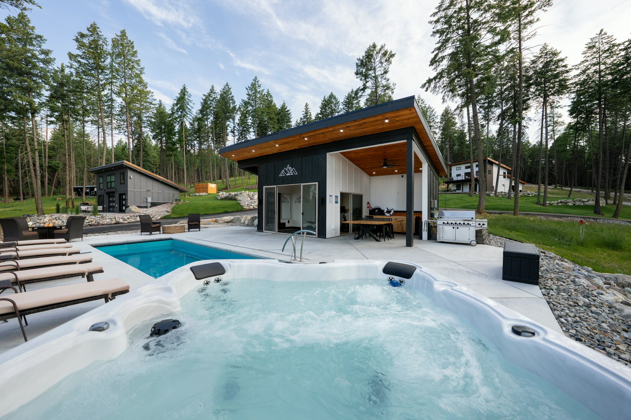 The estate hot tub in the foreground with the heated pool behind it, loungers alongside and the pool house's covered kitchen and dining