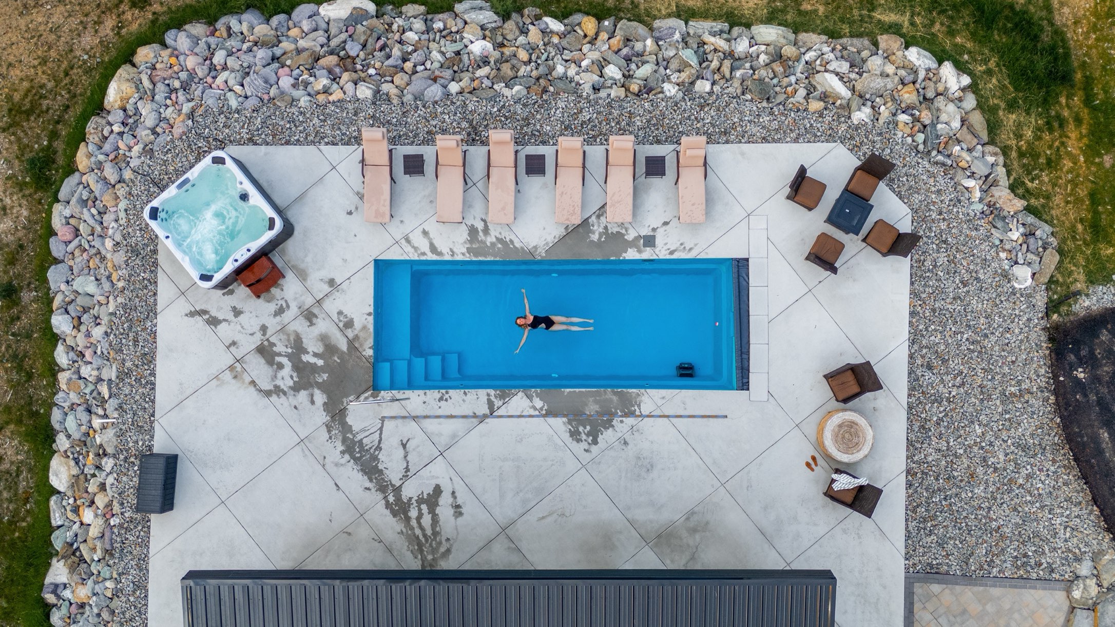 The estate pool from above with a swimmer floating in it, the hot tub, the loungers and the fire pit seating on the deck