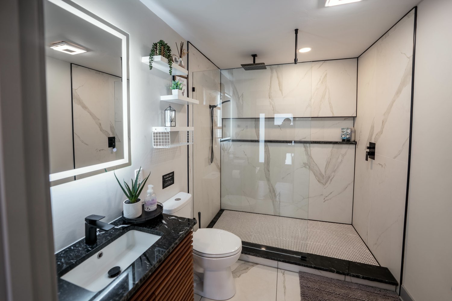 Bathroom at The Glacier House with a walk-in glass shower, marble-look tile, a black stone vanity top and a backlit mirror
