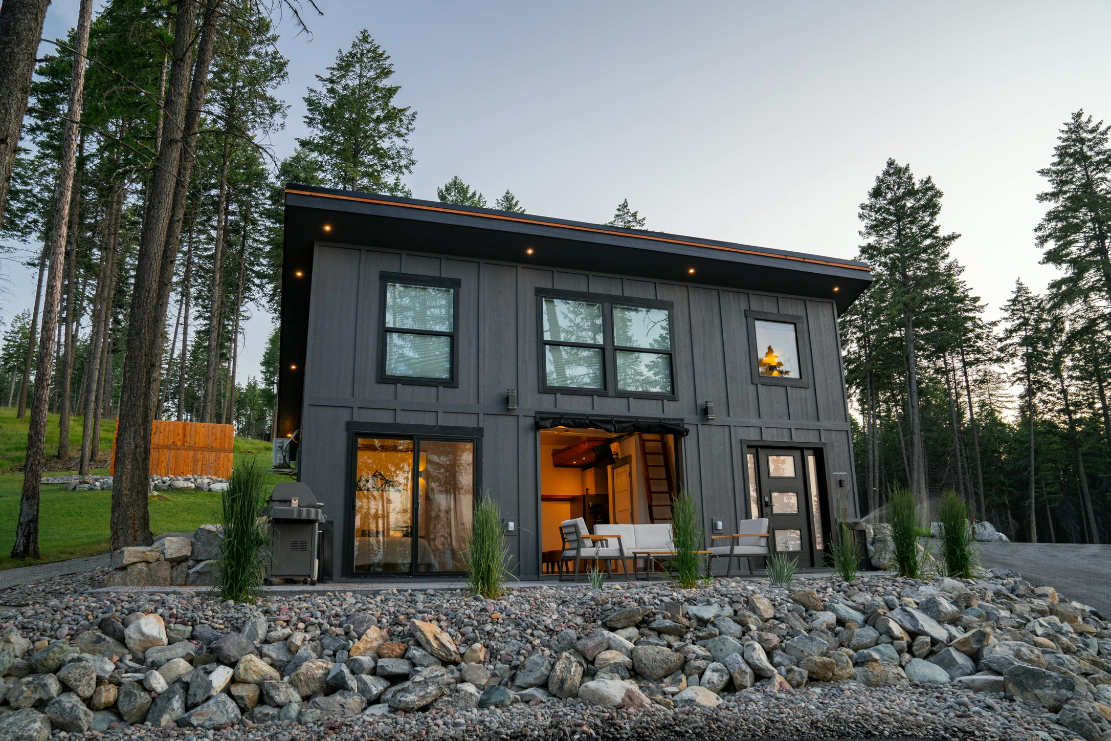 The Glacier House at dusk with its garage-door wall open onto a warmly lit interior and patio seating outside