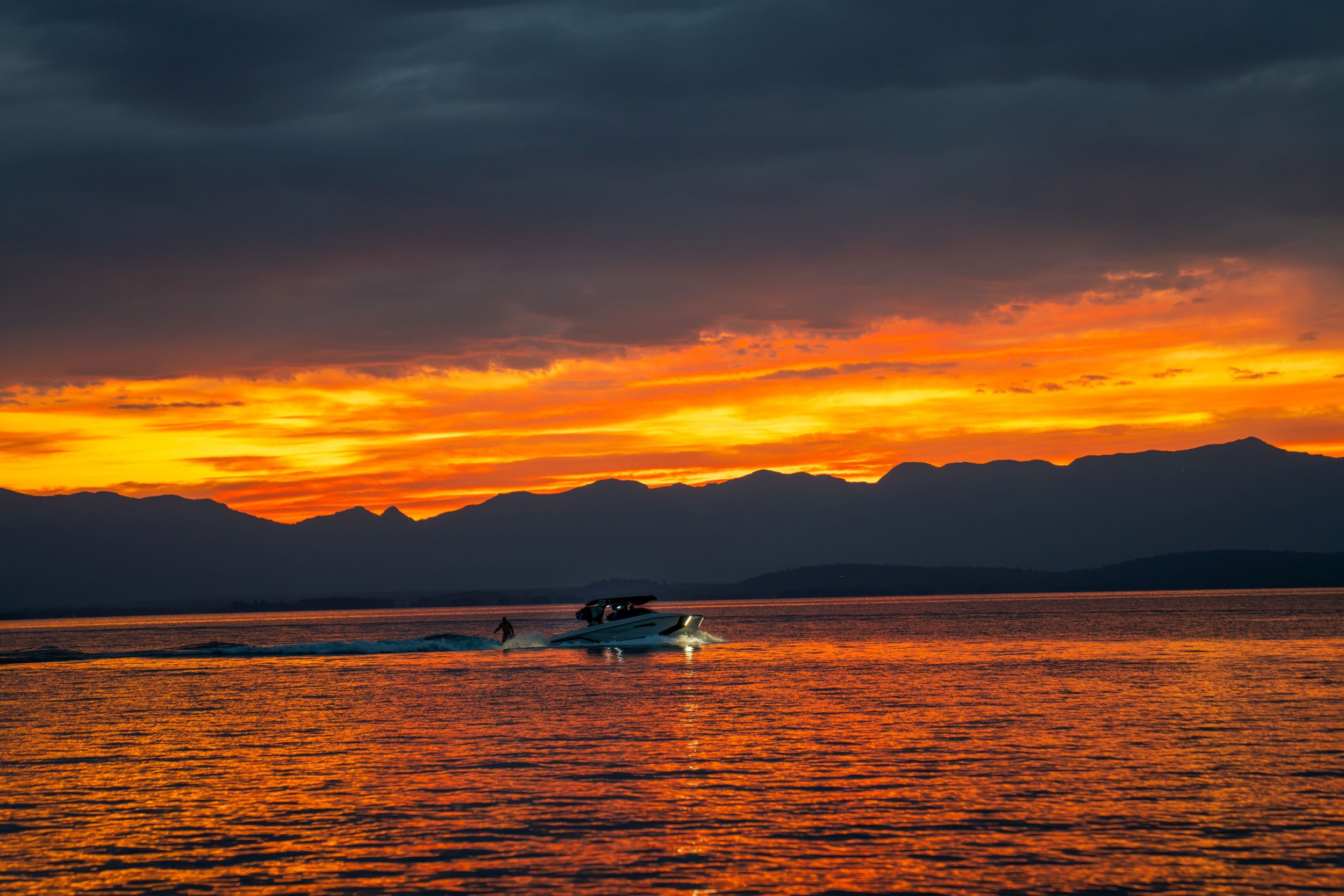 A boat crosses Flathead Lake beneath an orange sunset and mountain silhouettes