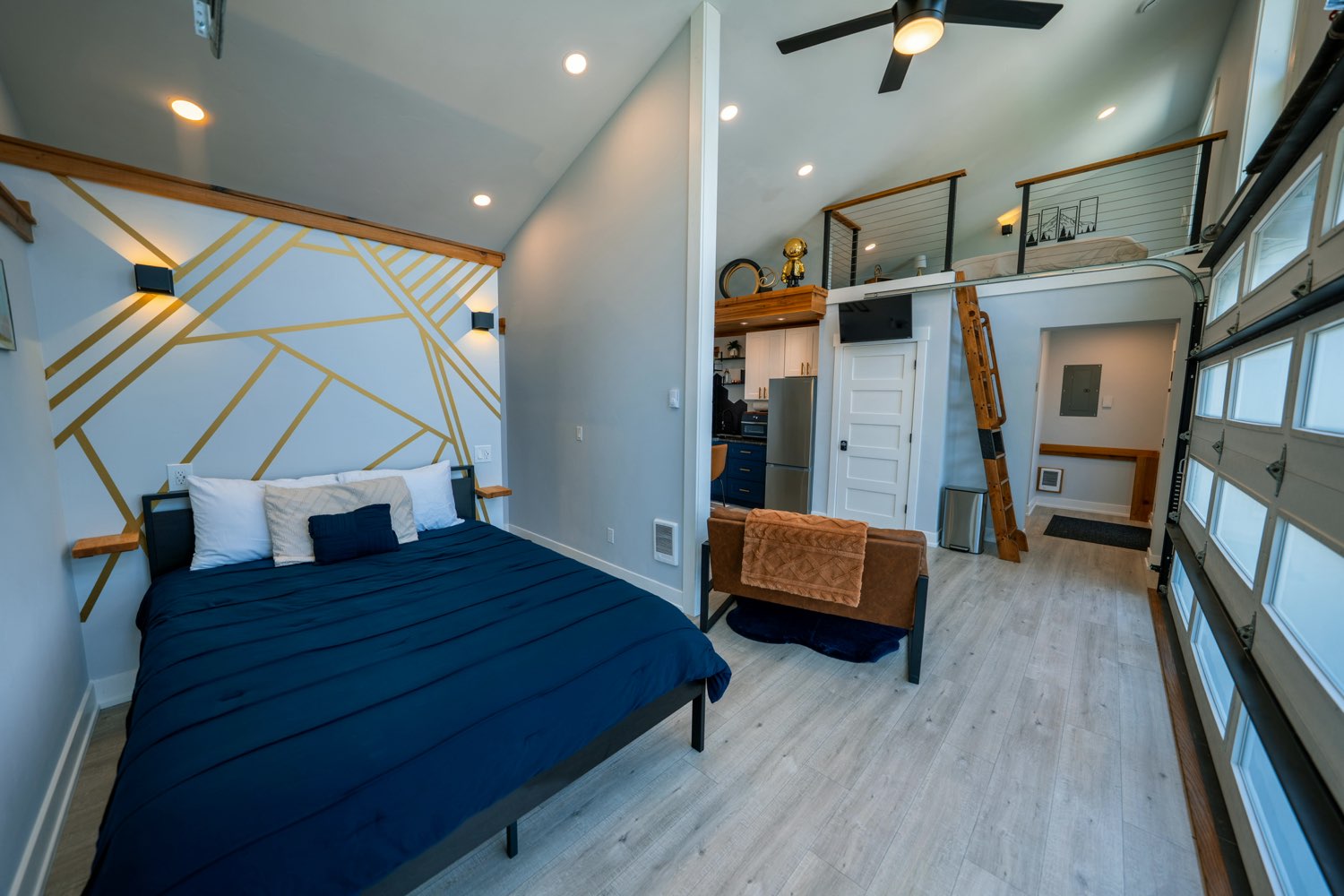 A bed beneath the geometric gold accent wall at The Lakeside House, with the loft above and the glass garage door alongside