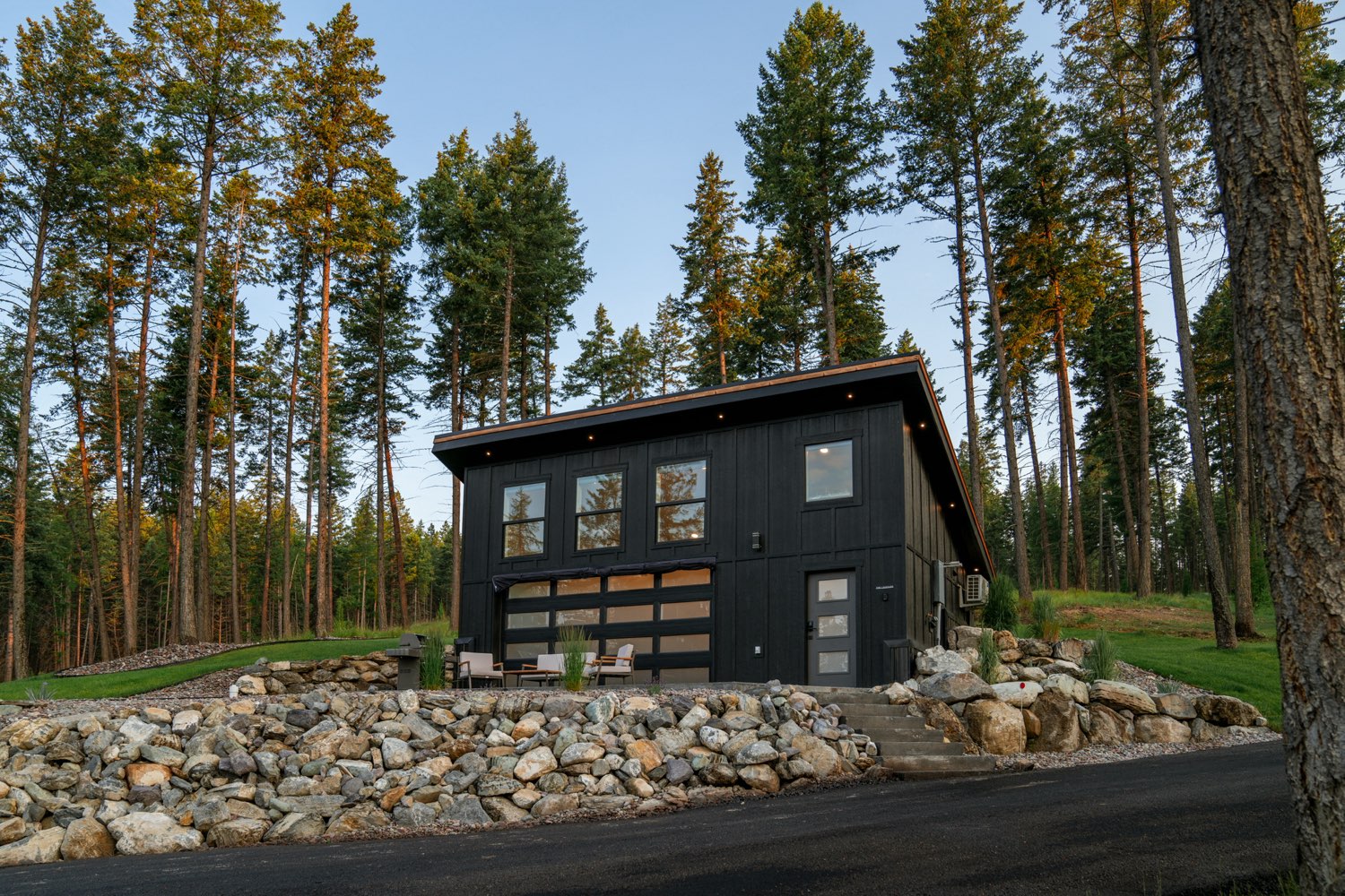 The Lakeside House, a black board-and-batten home with a glass garage door, set among tall pines above a boulder wall