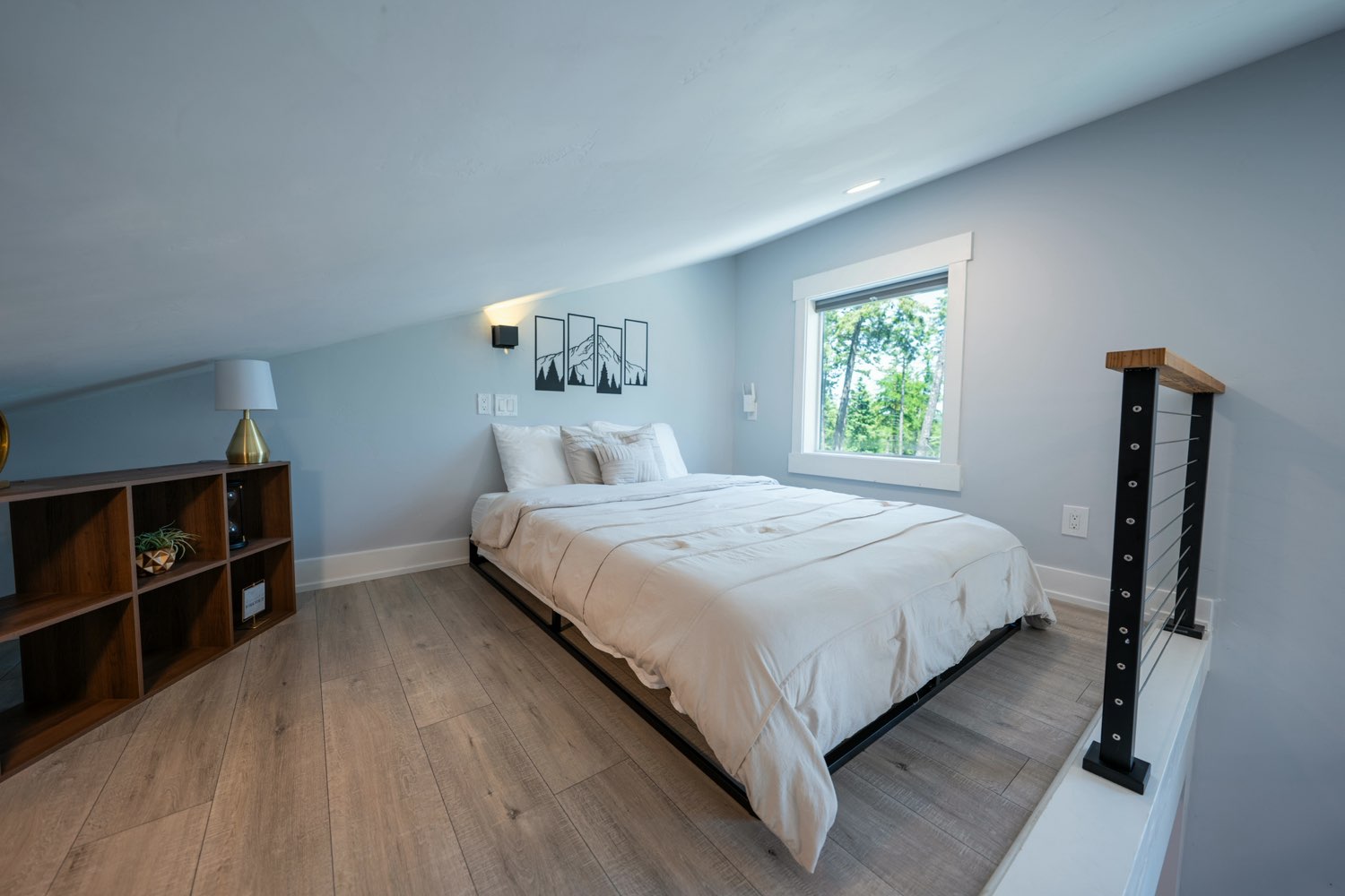 Loft bedroom at The Lakeside House under the sloped ceiling, with a window onto the pines and cable railing along the loft edge