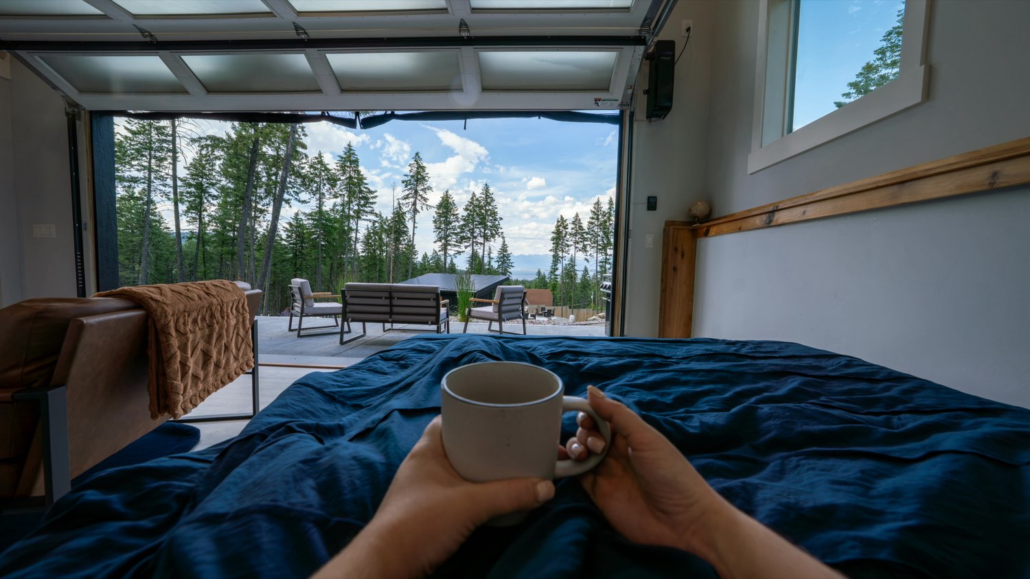 Hands holding a coffee mug in bed at The Lakeside House, with the glass garage door rolled up to the patio and the pines beyond