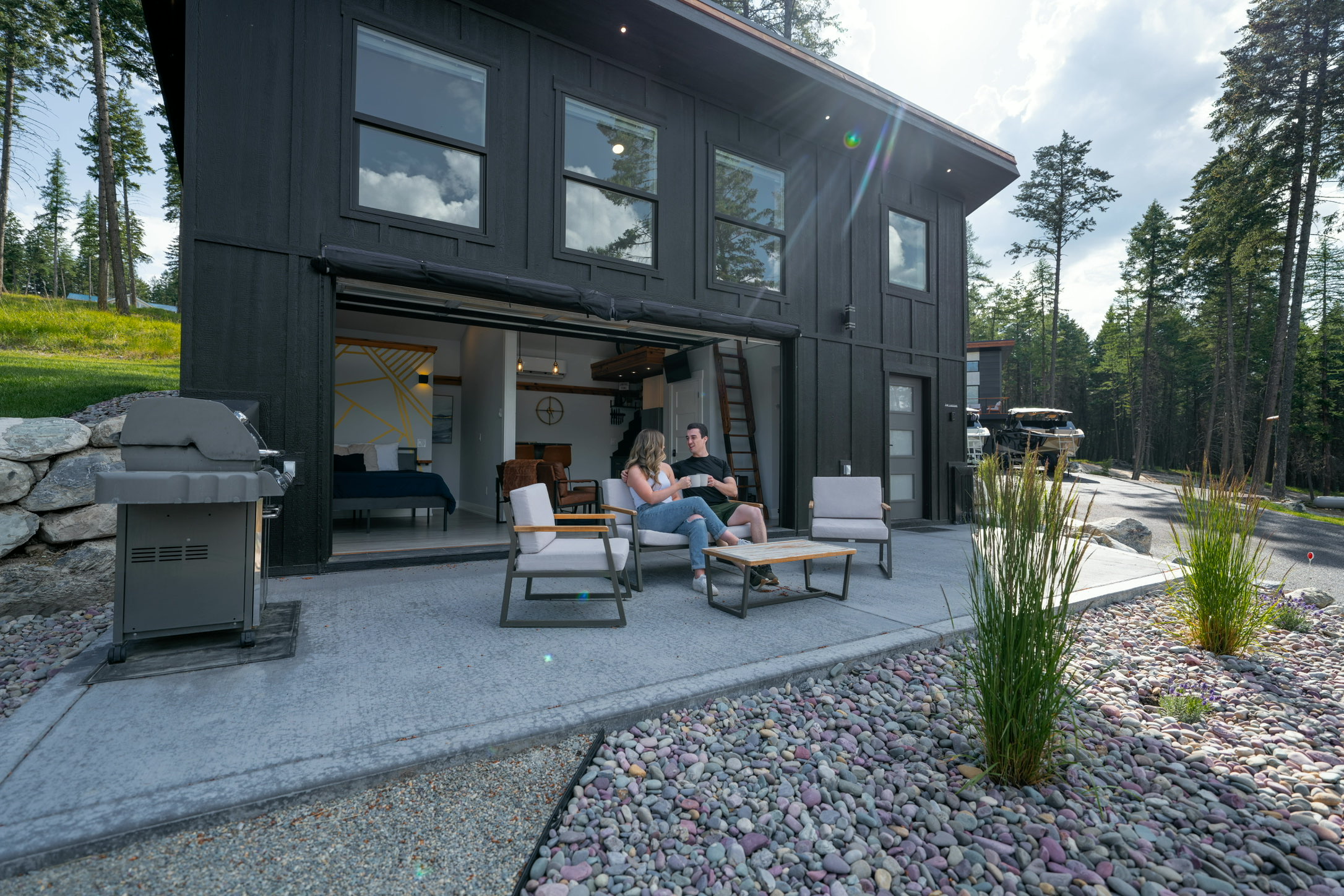 Two guests on the patio at The Lakeside House with the glass garage door rolled up to the living space and a gas grill alongside