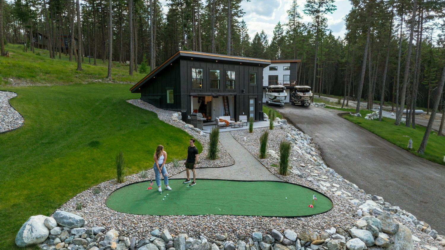 Two guests putting on the estate putting green below The Lakeside House, seen from above with the lawn and the forest around them
